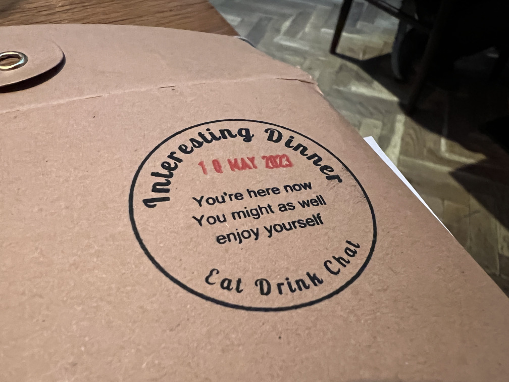 A circular ink stamp on a brown paper envelope: “Interesting Dinner / 18 May 2023 / You’re here now / You might as well enjoy yourself”